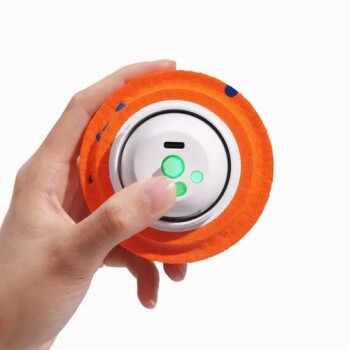 Hand hält ein orangefarbenes Stoff-Haustierspielzeug mit weißem elektronischem Zentrum und drei leuchtend grünen, pfotenförmigen LEDs.