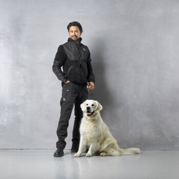 Eine Person in einer schwarzen und grauen Jacke steht neben einem lächelnden Golden Retriever vor einem grauen Hintergrund.