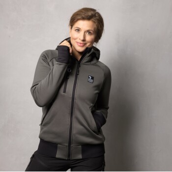 Eine Person, die einen dunkelgrauen Reißverschluss-Hoodie mit einem Logo trägt, das "DOG COACH" auf der linken Brust liest, vor einem hellgrauen Hintergrund steht.