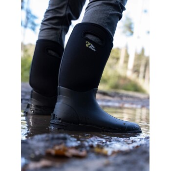 Eine Person, die schwarze wasserdichte Stiefel trägt, steht im flachen Wasser, mit einem verschwommenen Waldhintergrund. Die Stiefel haben ein Logo, das "Two Tracks" liest.
