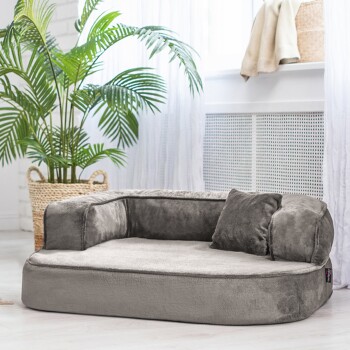 Ein plüschiges graues Haustierbett mit niedrigem Profil und einem kleinen Kissen, in einem hellen Raum mit einer Topfpflanze im Hintergrund.