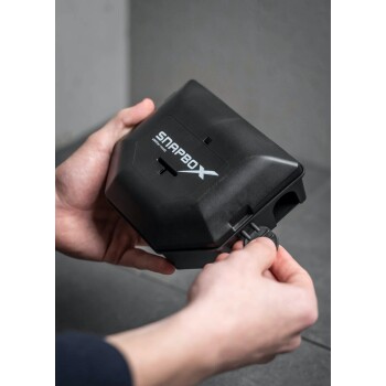 Eine Person hält ein schwarzes SNAPBOX-Haustierprodukt, das einen Schlitz und einen abnehmbaren Deckel aufweist, vor einem grauen Hintergrund.