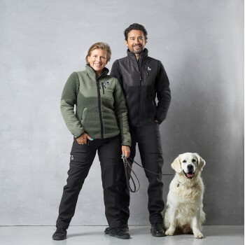Ein Mann und eine Frau stehen nebeneinander, beide tragen Outdoor-Jacken und Hosen, mit einem Golden Retriever, der neben ihnen sitzt.