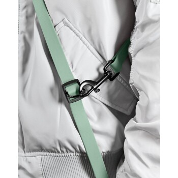 Eine Person in einer hellgrauen Jacke hält eine mintgrüne Hundeleine mit einem schwarzen Clip und steht vor einem schlichten weißen Hintergrund.