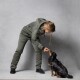 Eine Person in einer grünen Jacke und Hose füttert einen kleinen schwarz-braunen Hund mit einem Leckerli, während sie vor einem grauen Hintergrund steht.