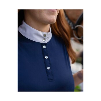 Nahaufnahme einer Frau, die ein marineblaues Shirt mit weißem Kragen und dekorativen Knöpfen trägt, lächelnd mit einem Pferd im Hintergrund.