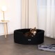 Ein kleiner brauner Hund sitzt bequem in einem schwarzen Hundebett, mit einer modernen Lampe und Vorhängen im Hintergrund.