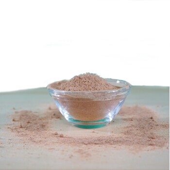 Eine klare Glasschüssel gefüllt mit einem feinen, hellbraunen Pulver, umgeben von einem kleinen Haufen des gleichen Pulvers auf einer hellen Oberfläche.