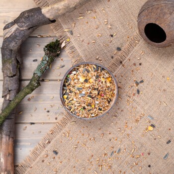 Eine Holzschüssel, gefüllt mit einer bunten Mischung aus Vogelfutter, steht auf einer Juteschicht, umgeben von verstreutem Futter und natürlichen Zweigen.