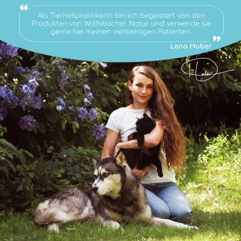 Eine Frau mit einer schwarzen Katze sitzt neben einem großen Hund in einem Garten mit lila Blumen. Der Text darüber bewirbt Wolfsbacher Naturprodukte.
