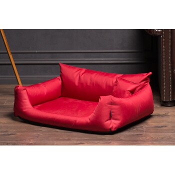 Rotes Haustierbett mit plüschigen Seiten und einem weichen Boden, auf Holzboden neben einem braunen Ledersessel platziert.