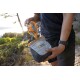 Eine Person hält einen grauen und orangefarbenen Ruffwear-Beutel, der teilweise geöffnet ist, mit einer malerischen Outdoor-Hintergrundszene aus Grün und Wasser.