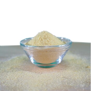 Eine klare Glasschüssel gefüllt mit einem feinen, gelblichen Pulver, umgeben von verstreutem Pulver auf einer hellen Oberfläche.