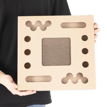 Eine Person, die einen quadratischen Papp-Kratzbaum für Katzen mit ausgeschnittenen Formen und einer strukturierten Mitte hält, entworfen für das Spielen und Kratzen von Haustieren.