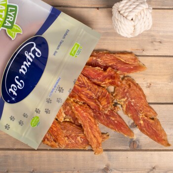 Eine Tüte mit Lyra Pet natürlichen Hundeleckerlis ist teilweise geöffnet und zeigt Streifen von getrocknetem Fleisch auf einer Holzoberfläche, mit einem weißen Seilspielzeug in der Nähe.