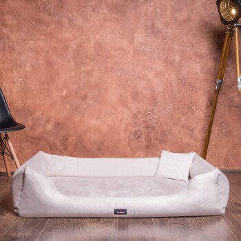 Ein plüschiges, cremefarbenes Haustierbett mit erhöhten Rändern und einem kleinen Kissen, vor einer strukturierten braunen Wand und einem Holzboden.