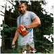 Ein Mann in einem grauen T-Shirt hält eine Leine und greift in eine orangefarbene Haustiertasche, während ein brauner Hund zu ihm in einem Wald aufblickt.