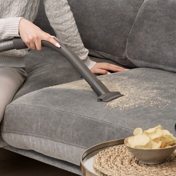 Eine Person, die einen Staubsaugeraufsatz verwendet, um Krümel von einer grauen Couch zu reinigen, mit einer Schüssel Kartoffelchips auf einem nahegelegenen Tisch.