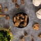 Eine schwarze Schüssel mit getrockneten Haustierleckereien auf einer grauen Oberfläche, umgeben von verstreuten Leckereien und einem geflochtenen Korb mit grünem Gras.