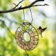 Ein Vogelfutterspender in Ringform, gefüllt mit Saatkuchen, hängt von einem Baumast, während ein kleiner Vogel in der Nähe fliegt.