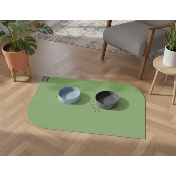 Haustier-Futterstation mit blauen und grauen Schüsseln auf einer grünen Matte, auf Holzboden neben Pflanzen und Stuhl platziert.