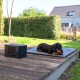 Ein Dackel liegt auf einer grauen Matte neben einem kleinen schwarzen Gerät mit einem blauen Netzschalter, das auf einer Holzterrasse in einem Garten steht.