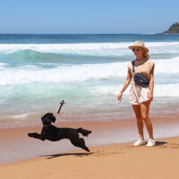 Eine Frau in einem beigen Outfit und Hut steht am Strand und beobachtet einen schwarzen Hund, der fröhlich mit einem Stock im Mund springt, mit Wellen im Hintergrund.