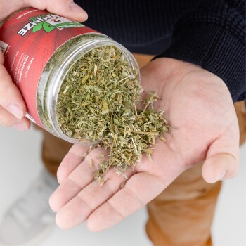 Eine Person gießt getrocknete Kräuter aus einem klaren Glasbehälter in ihre Handfläche und zeigt den grünen, blättrigen Inhalt. Der Behälter hat ein rotes Etikett.