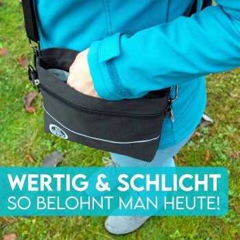 Eine Person in einer blauen Jacke greift in eine schwarze, mit Reißverschluss verschlossene Tasche für Haustierleckerlis gegen Gras. Text: "WERTIG & SCHLICHT SO BELOHNT MAN HEUTE!"