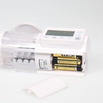 Ein weißer elektronischer Futterspender für Haustiere mit einem digitalen Display und Steuerknöpfen, der ein Batteriefach mit zwei AA-Batterien zeigt.