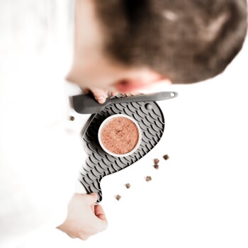 Eine Hand hält eine schwarze, fischförmige Futterschüssel für Haustiere, die mit Nassfutter für Katzen gefüllt ist, mit kleinen Futterstücken, die in der Nähe verstreut sind.