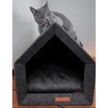 Eine graue Katze sitzt auf einem grauen Filzhaustierhaus mit einem schwarzen Kissen und leckt sich die Pfote. Das Haus hat ein dreieckiges Dach und ein Logo-Tag.