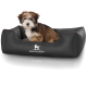 Ein kleiner, flauschiger Hund sitzt bequem in einem schwarzen Haustierbett mit einem gesteppten Innenraum, das das Logo 'KNUFFELWUFF' zeigt.