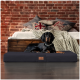 Ein schwarzer Dobermann liegt auf einem stilvollen Hundebett in einem gemütlichen Raum mit einer Ziegelwand, umgeben von dekorativen Kissen und einer gestrickten Decke.