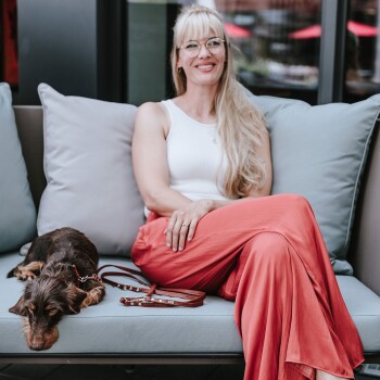 Eine Frau in einem weißen Tanktop und korallenfarbenen Hosen sitzt auf einer Couch mit einem kleinen braunen Hund, der neben ihr ruht, eine Leine in der Nähe.