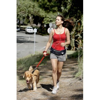 Eine Frau in einem roten Tanktop und grauen Shorts joggt auf einem Weg mit einem Golden Retriever an einer roten Leine, umgeben von Grün.