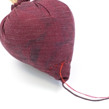 Ein plüschiges, herzförmiges Haustierspielzeug aus tiefburgunderfarbener Stoff mit sichtbaren Nähten und einem Kordelzugverschluss an der Oberseite.