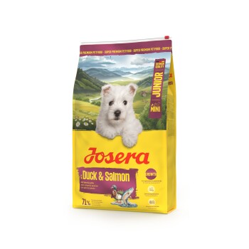 Josera Junior Mini Ente & Lachs Hundefutterbeutel mit weißem Welpen und malerischem Bergpanorama im Hintergrund.