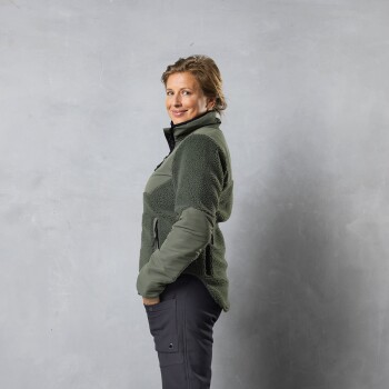 Eine Person in einer grünen Fleecejacke mit einem Reißverschlusskragen und schwarzen Hosen, vor einem grauen Hintergrund stehend.
