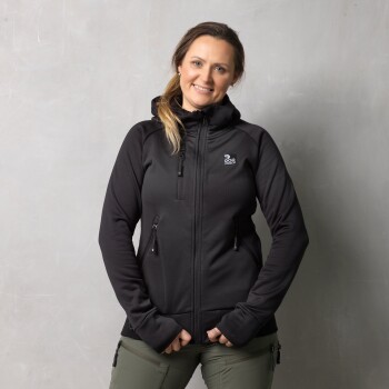 Eine Person trägt eine schwarze Zip-Jacke mit einem Logo auf der linken Brust, steht vor einem grauen Hintergrund.