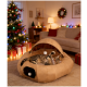 Getigerte Katze liegt in einem hellbraunen, höhlenartigen Katzenbett mit hängendem Pompon-Spielzeug; Weihnachtsbaum, Geschenke und gemütliche Wohnzimmerdekoration im Hintergrund.