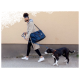 Eine Person führt einen schwarz-weißen Hund an der Leine, trägt eine große blaue Haustiertasche über der Schulter, vor einer beigen Wand.