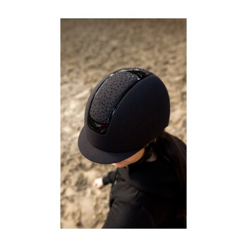 Eine Nahaufnahme eines Reiters, der einen eleganten schwarzen Reithelm mit belüftetem Design trägt, vor dem Hintergrund einer sandigen Reitarena.