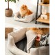 Ein flauschiger orangefarbener Pomeranian sitzt bequem in einem beigen Haustierbett mit einem weichen grauen Innenraum, neben einer Topfpflanze und einem Holzregal.