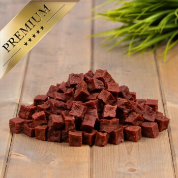 Ein Haufen kleiner, quadratischer, rötlich-brauner Haustier-Leckerlis auf einer Holzoberfläche, mit einer grünen Pflanze im Hintergrund und einem goldenen 'PREMIUM'-Abzeichen in der Ecke.