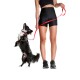 Eine Frau in schwarzen Sportshorts und einem Crop-Top hält eine rote Hundeleine, während ein kleiner schwarz-weißer Hund auf seinen Hinterbeinen steht und nach oben schaut.