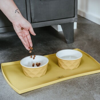 Eine Hand gießt Haustierfutter in eine gelbe, diamantmusterte Schüssel auf einer passenden Matte, neben einer Schüssel mit Wasser.