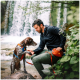 Eine Person kniet neben einem braun-weißen Hund mit einem blauen und orangefarbenen Geschirr, in der Nähe eines Wasserfalls und üppigem Grün.