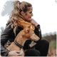 Eine Frau in einer schwarzen Lederjacke und gelbem Schal sitzt im Freien und hält sanft einen braunen Hund mit einem blauen Nietenhalsband.