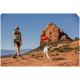 Eine Person in einem hellblauen Hemd und Shorts wirft einen roten Frisbee, während ein weißer Hund in einer felsigen Landschaft läuft, um ihn zu fangen.